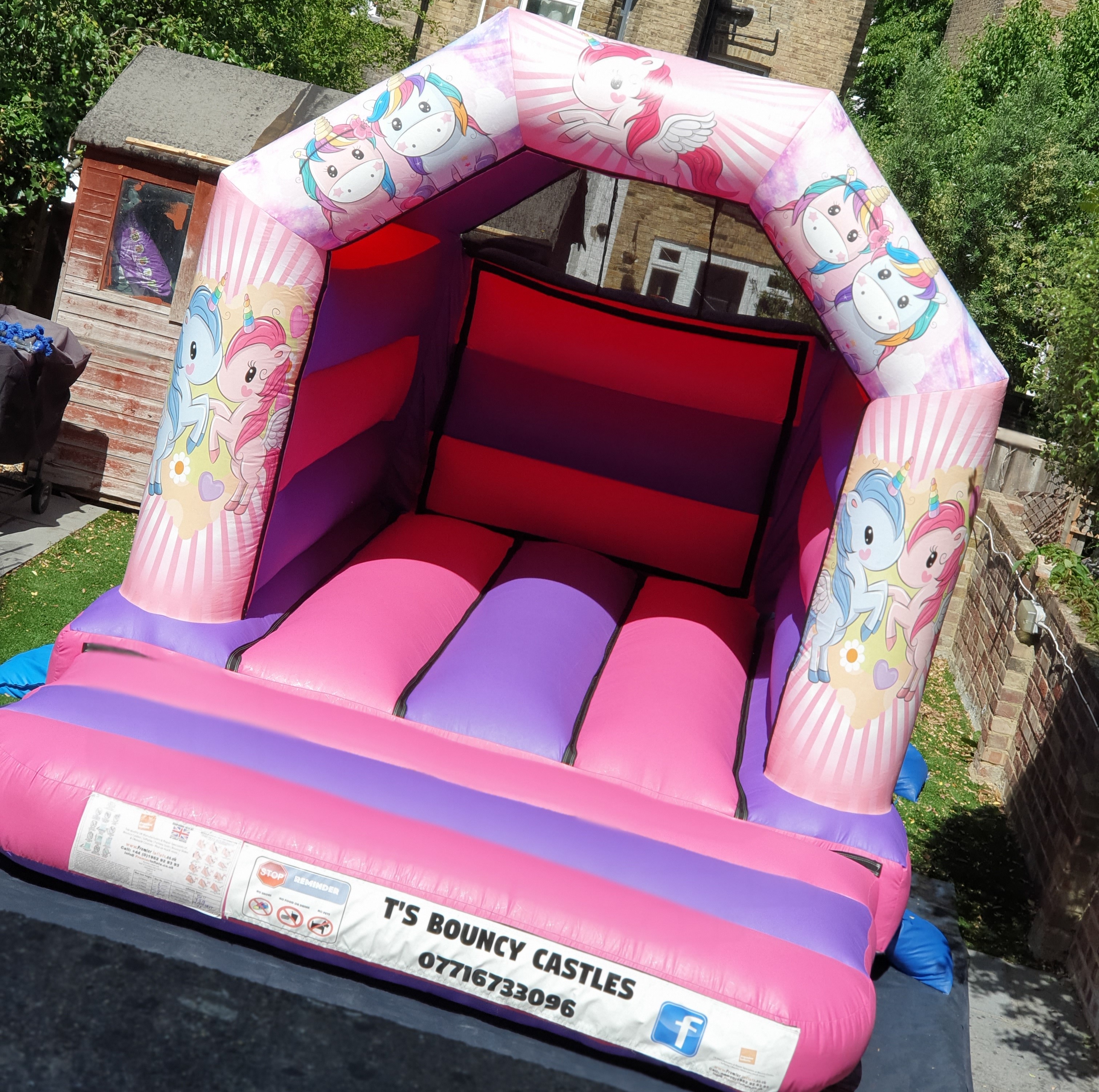 Pink and purple inflatable bounce castle with unicorn designs on sides, red interior climbing wall and padded floor in a residential garden